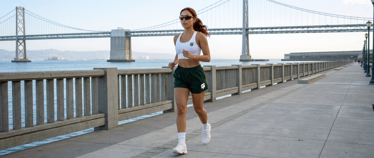 A woman jogging on an outdoor waterfront path with a large suspension bridge and water in the background. She is wearing a white sports bra, dark green athletic shorts, white socks, and white running shoes.