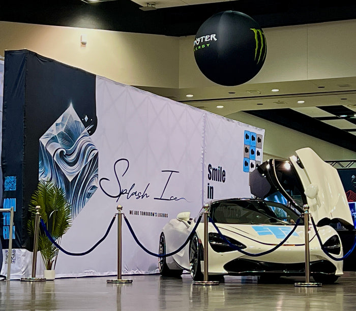 A white and black McLaren sports car with its gull-wing door open, displayed next to a large white and navy Splash Ice event backdrop with stanchion ropes in an indoor exhibition space.