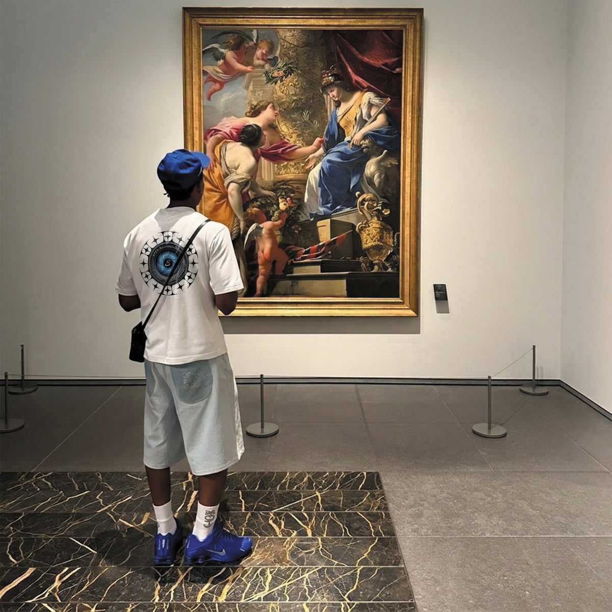 A man in a white t-shirt, light gray shorts, blue cap, and blue sneakers stands with his back to the camera, looking at a large framed classical painting hanging on a white gallery wall.